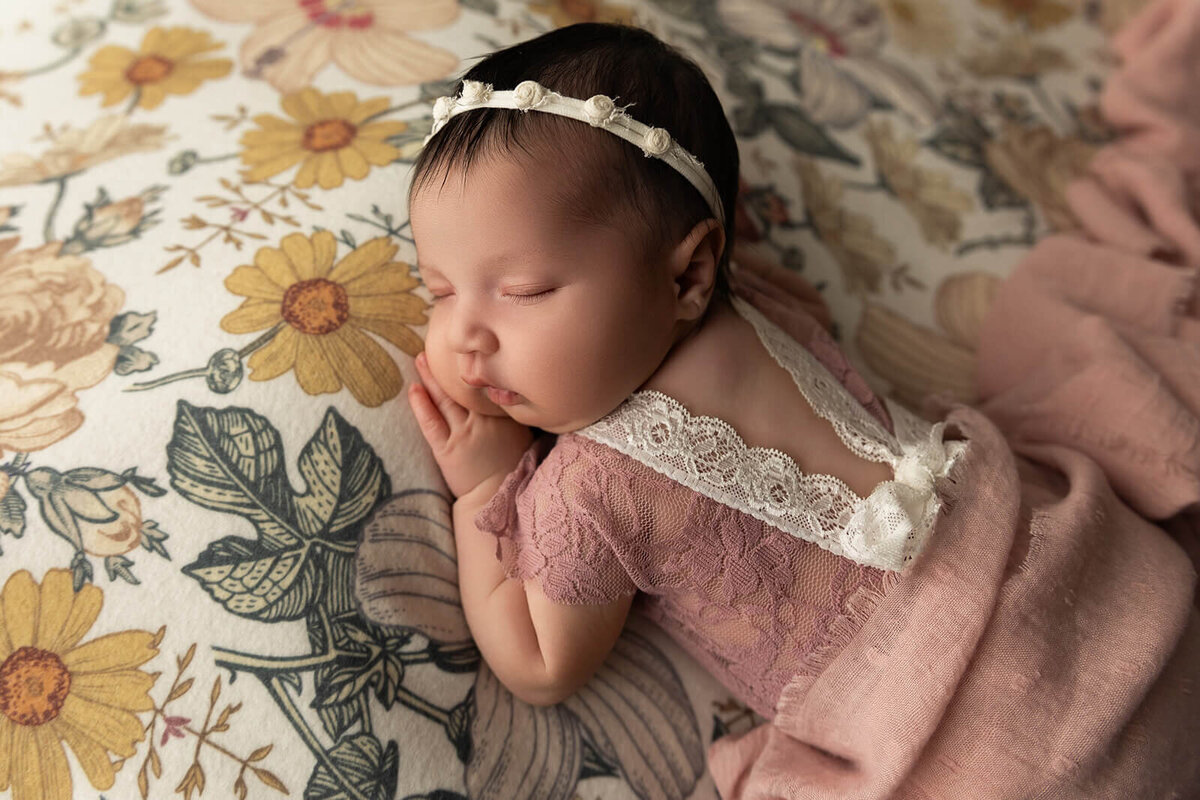 A peaceful newborn baby sleeps on a floral-patterned blanket, dressed in a pink lace-trimmed outfit. The baby’s head is turned, revealing soft features and a delicate headband. The setting creates a calm, serene atmosphere.