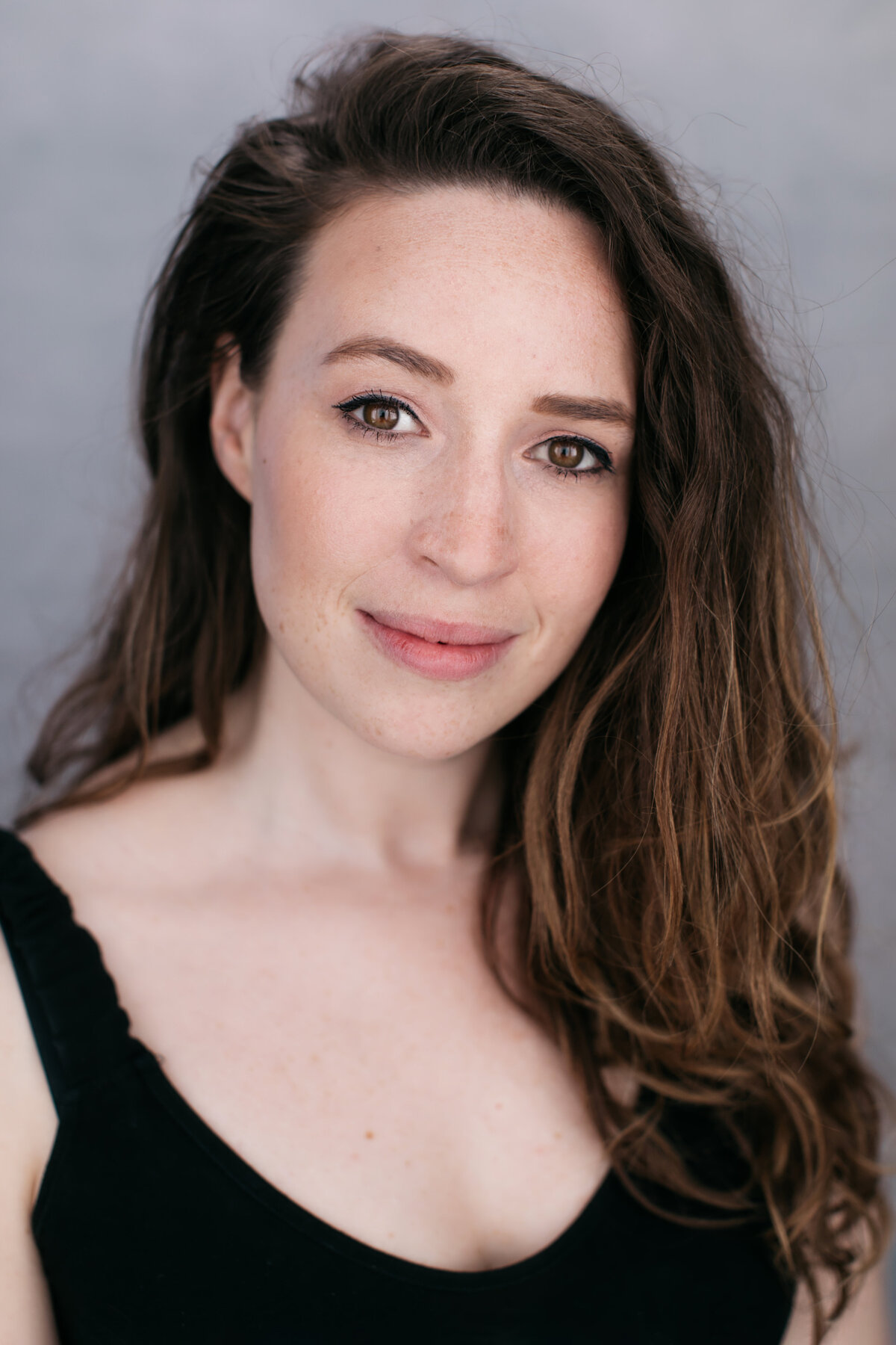 Castingfoto van Janis Van Dorsselaer. Vrouw met bruin haar, bruine ogen en zwarte tanktop voor een grijze achtergrond.