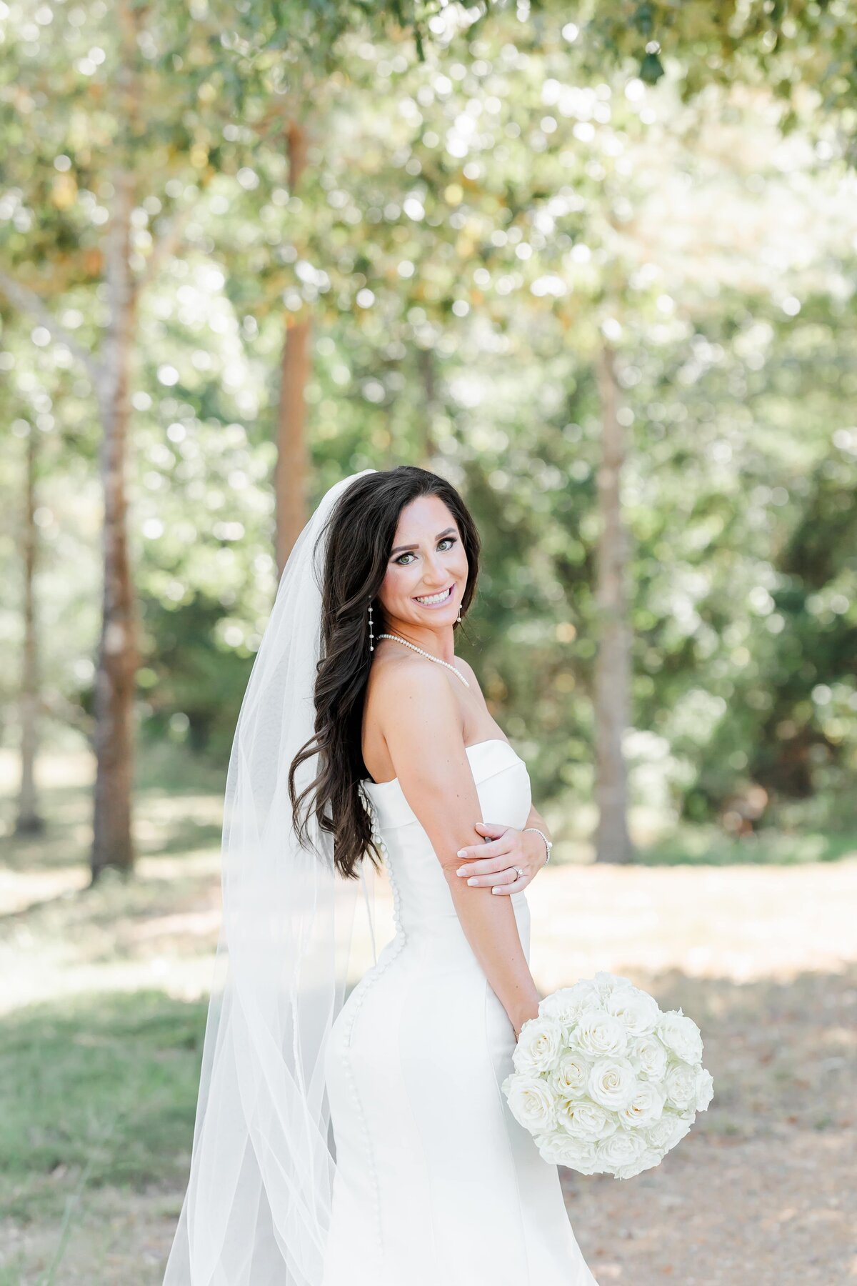 An image from an Arkansas wedding photographer of a bride standing smiling over her shoulder with her flowers outside the Venue at Oakdale.