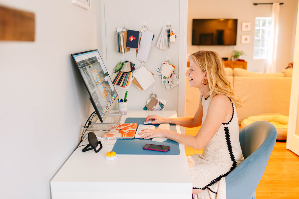 A woman with blonde hair and gold hoop earrings sits at a white desk, smiling while working on a design project on her computer. Swatches and color samples hang on the wall behind her, and a cozy living space is visible in the background.