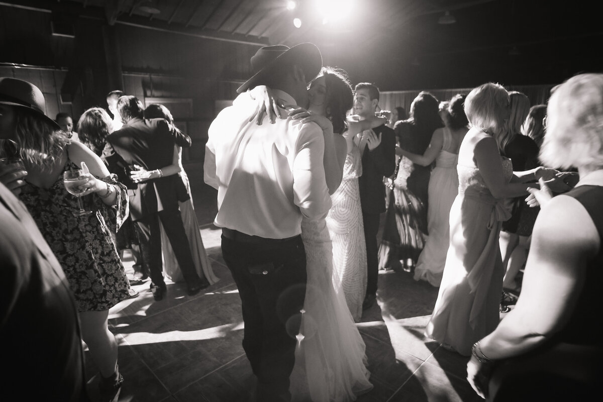 A bride and groom share a romantic first dance, with the groom wearing a cowboy hat, surrounded by guests on a lively wedding dance floor.