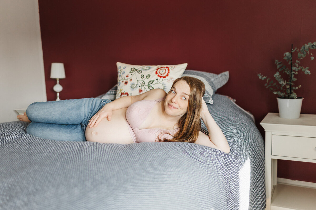 Maternity portrait of pregnant red headed woman wearing jeans and a pink bra laying sideways on her bed