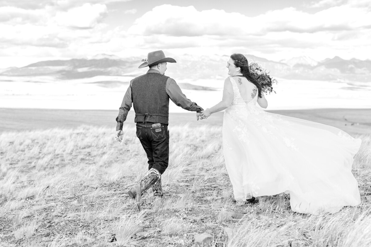 Bozeman, Montana wedding on a ranch in front of the Rocky mountains. Black and white photograph of bride and groom holding hands and walking away with bride's dress flowing in the wind