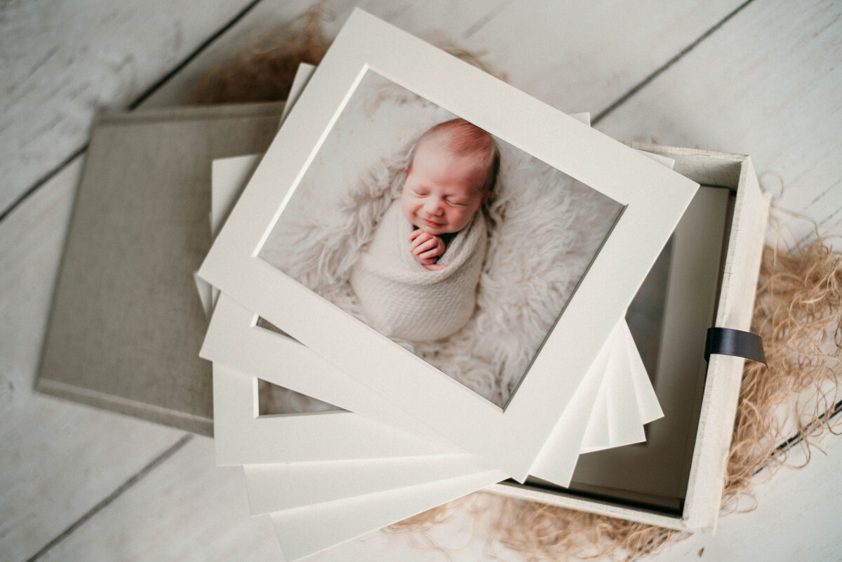 Printed picture of newborn baby in white swaddle. Print is matted and in a white box with other matted prints.