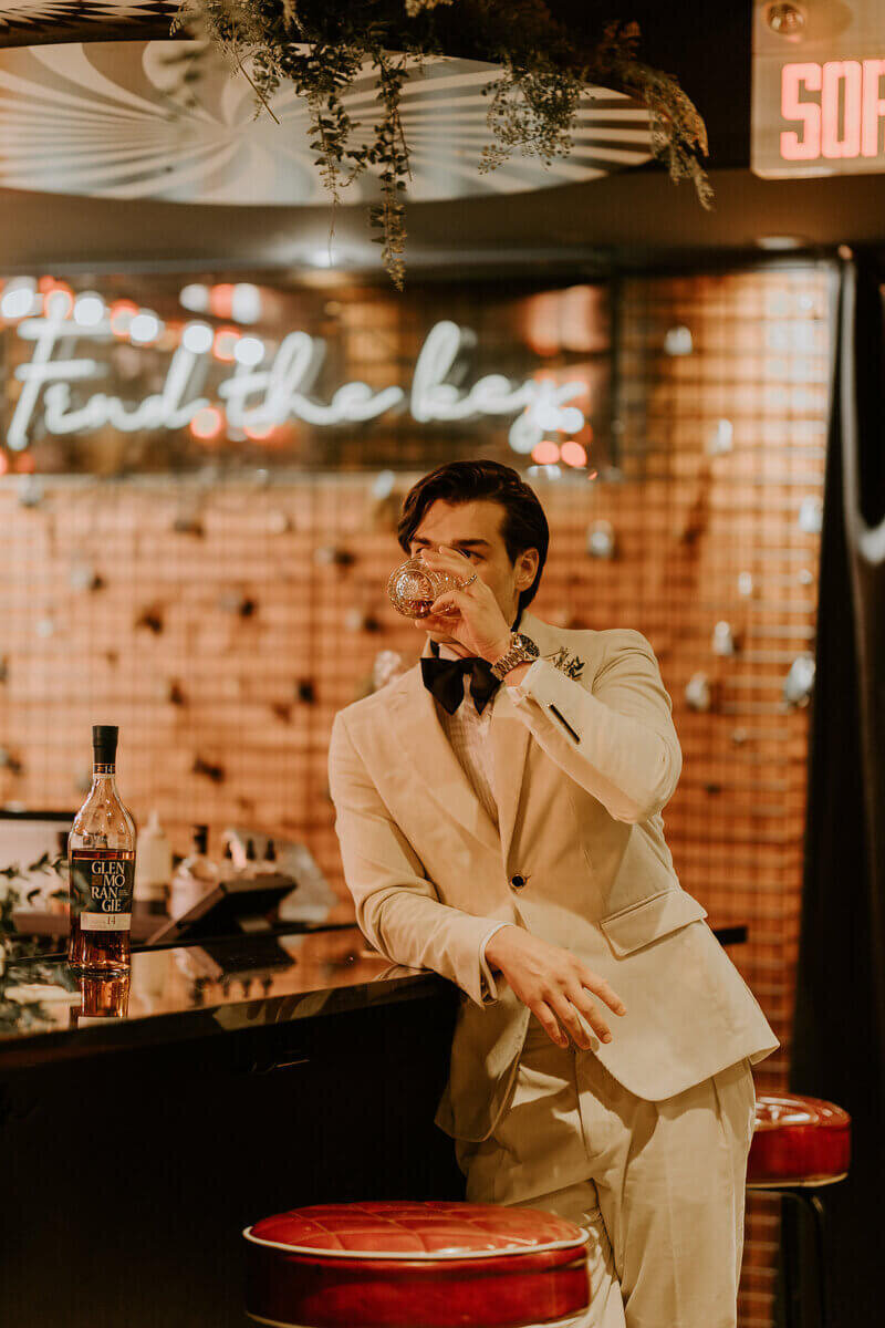 Marié en costume beige accoudé à un bar et buvant un verre d'alcool pour un shooting photo mariage. Décor vintage avec néon.