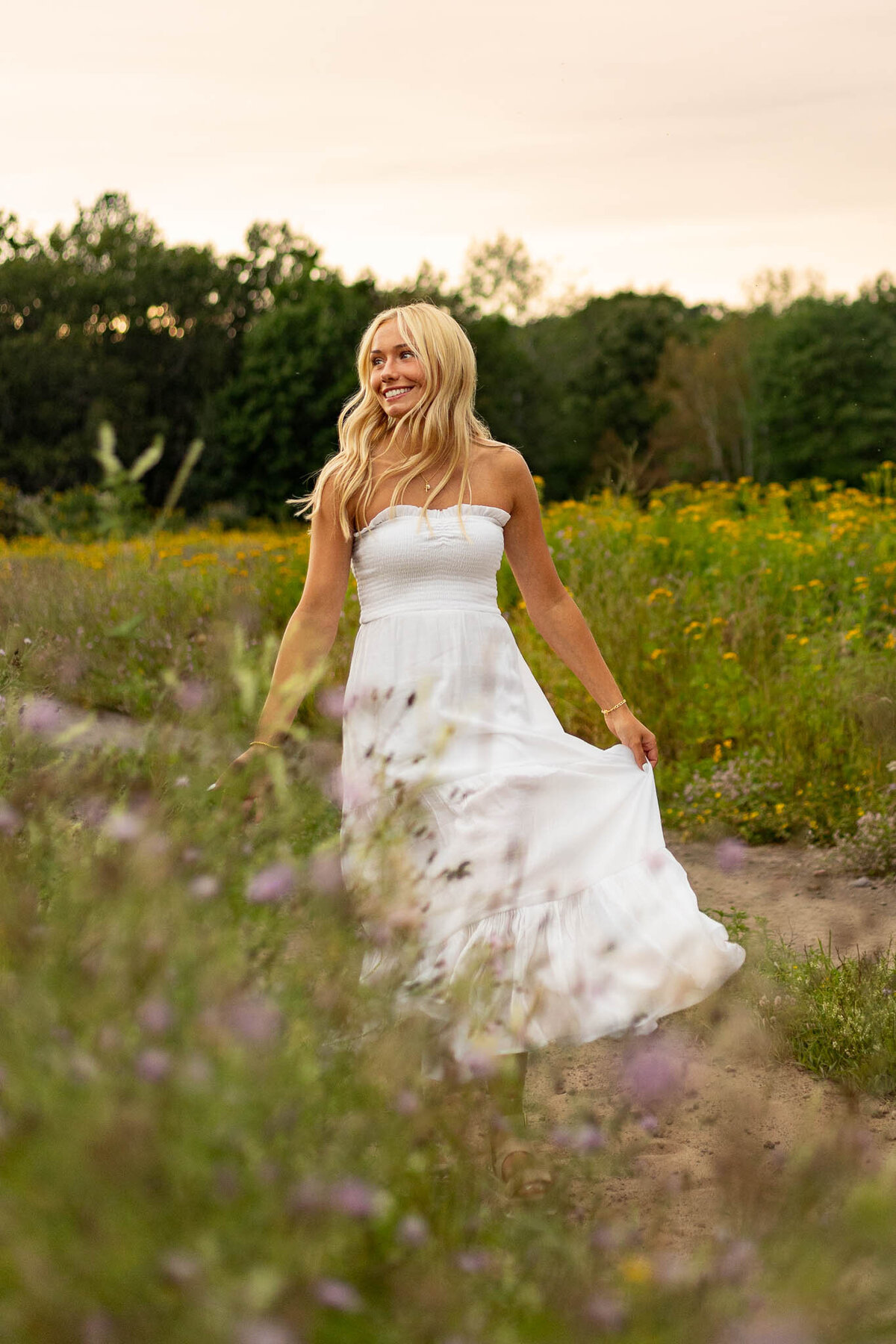 A senior girl wears a beautiful white gown in a scenic field, with golden sunlight casting a soft, ethereal glow for a bridal-inspired portrait.
