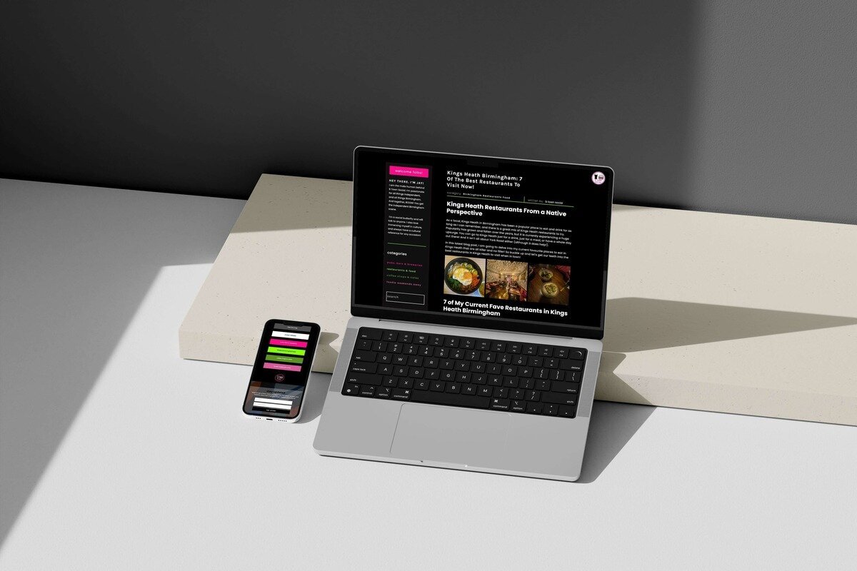 A laptop and a smartphone placed on a light beige surface, displaying the B Town Social food blog. The laptop screen shows an article titled "Kings Heath Birmingham: 7 Of The Best Restaurants To Visit Now!" with images of various dishes. The smartphone displays a colourful menu from the same website. The background features a minimalist setup with neutral tones and a grey wall, creating a clean and modern look