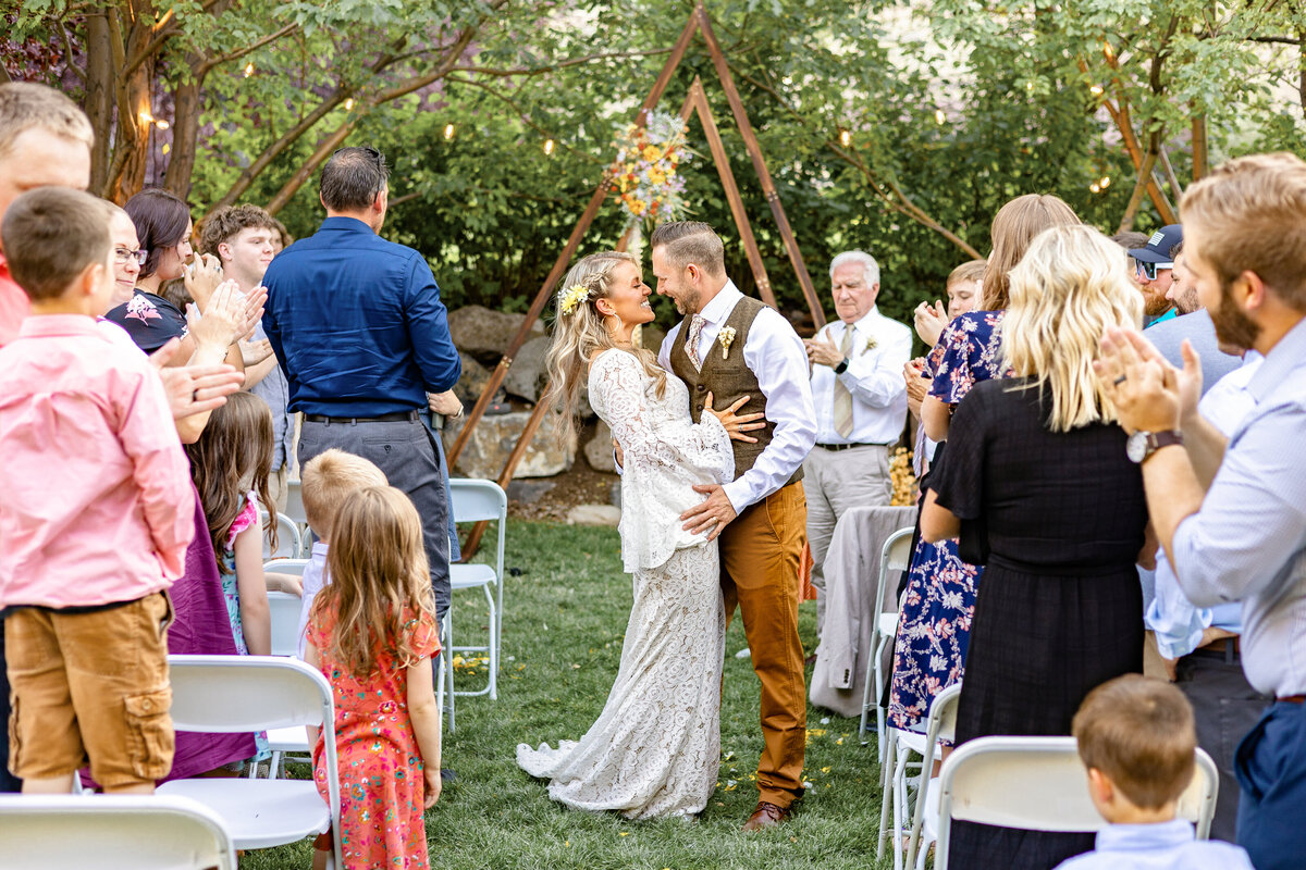 The bride and groom share a romantic kiss at the end of the ceremony as guests cheer and applaud around them in a beautiful outdoor setting in Provo Canyon.