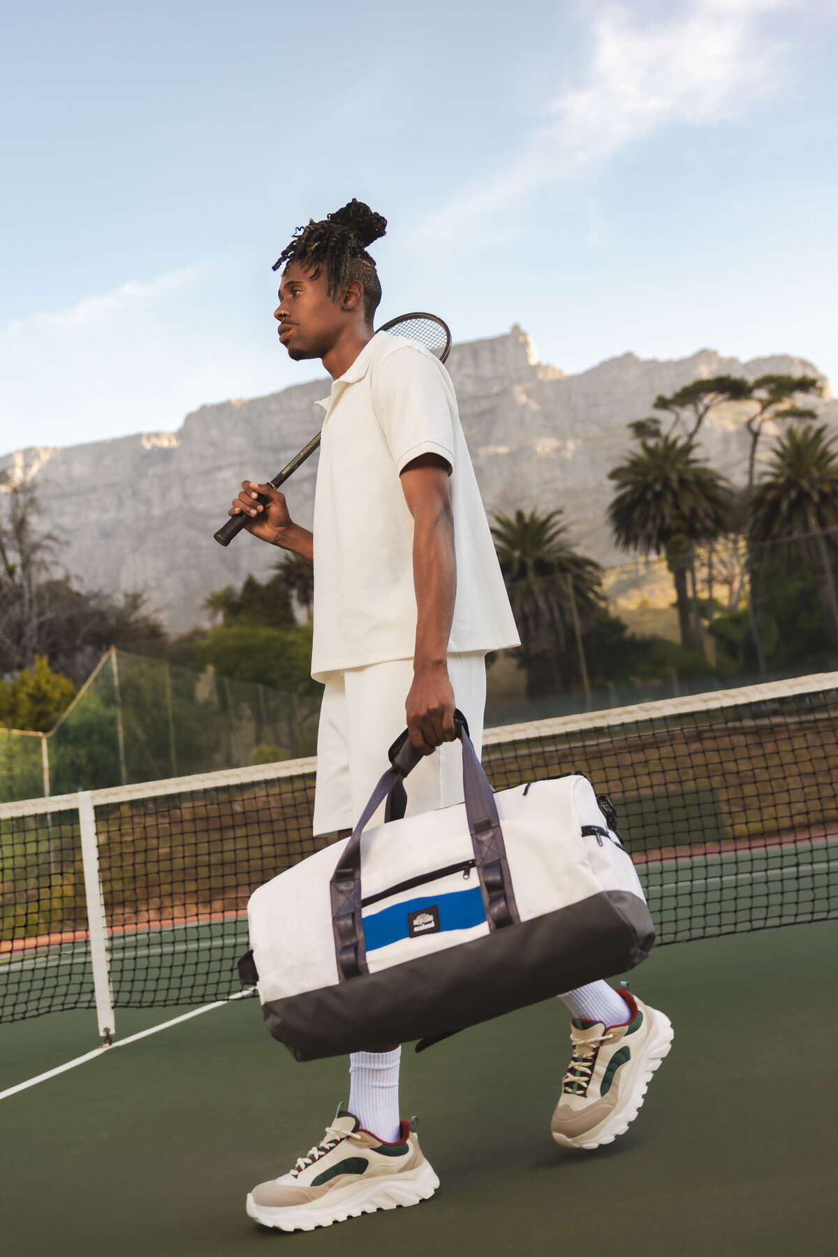 Stylish man in a white tennis outfit walking across the court with a racket over his shoulder and a large duffle bag in hand, with Table Mountain and palm trees in the background.