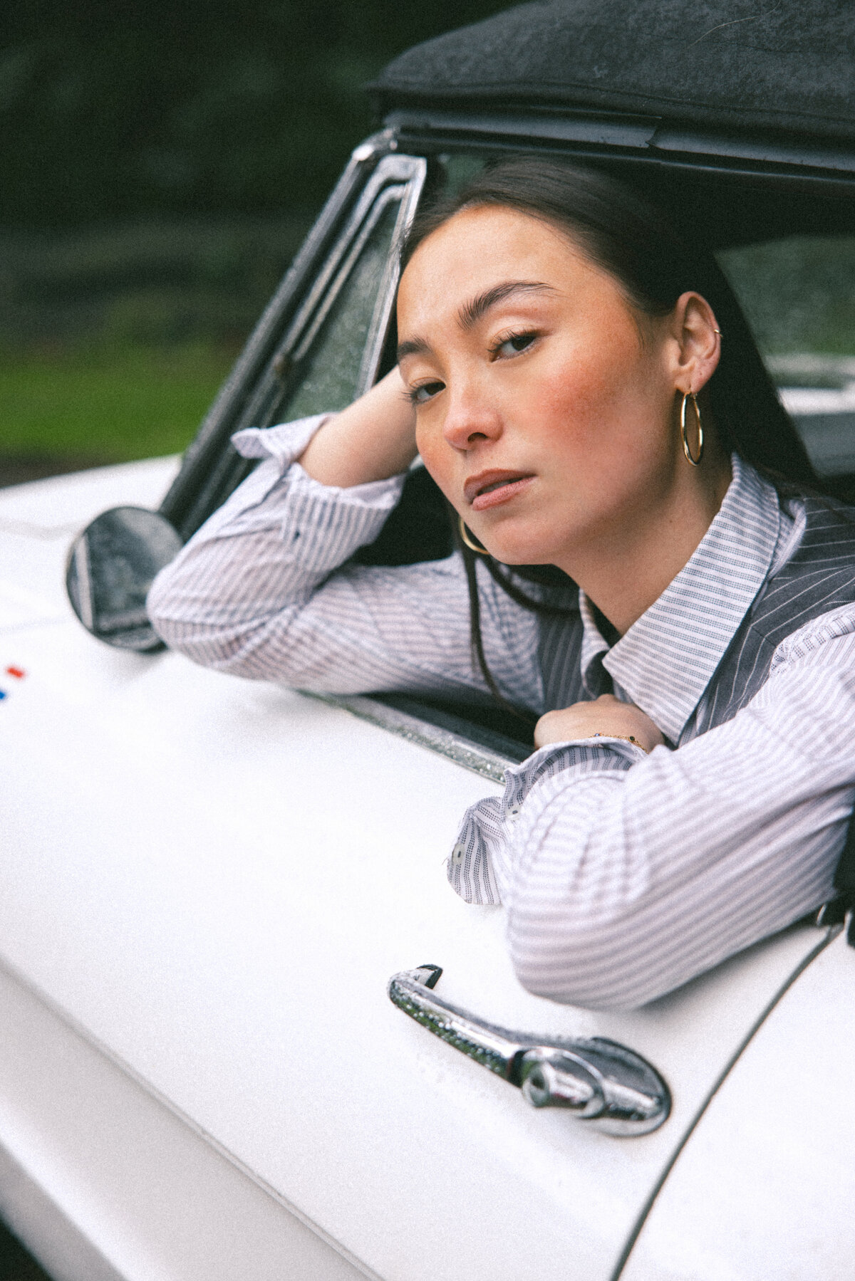Capturing a moment of calm and reflection, this image shows a woman leaning out of the window of a white vintage car, her expression contemplative. The light softly highlights her features, creating a serene and introspective mood, photographed by Bo Eppink.