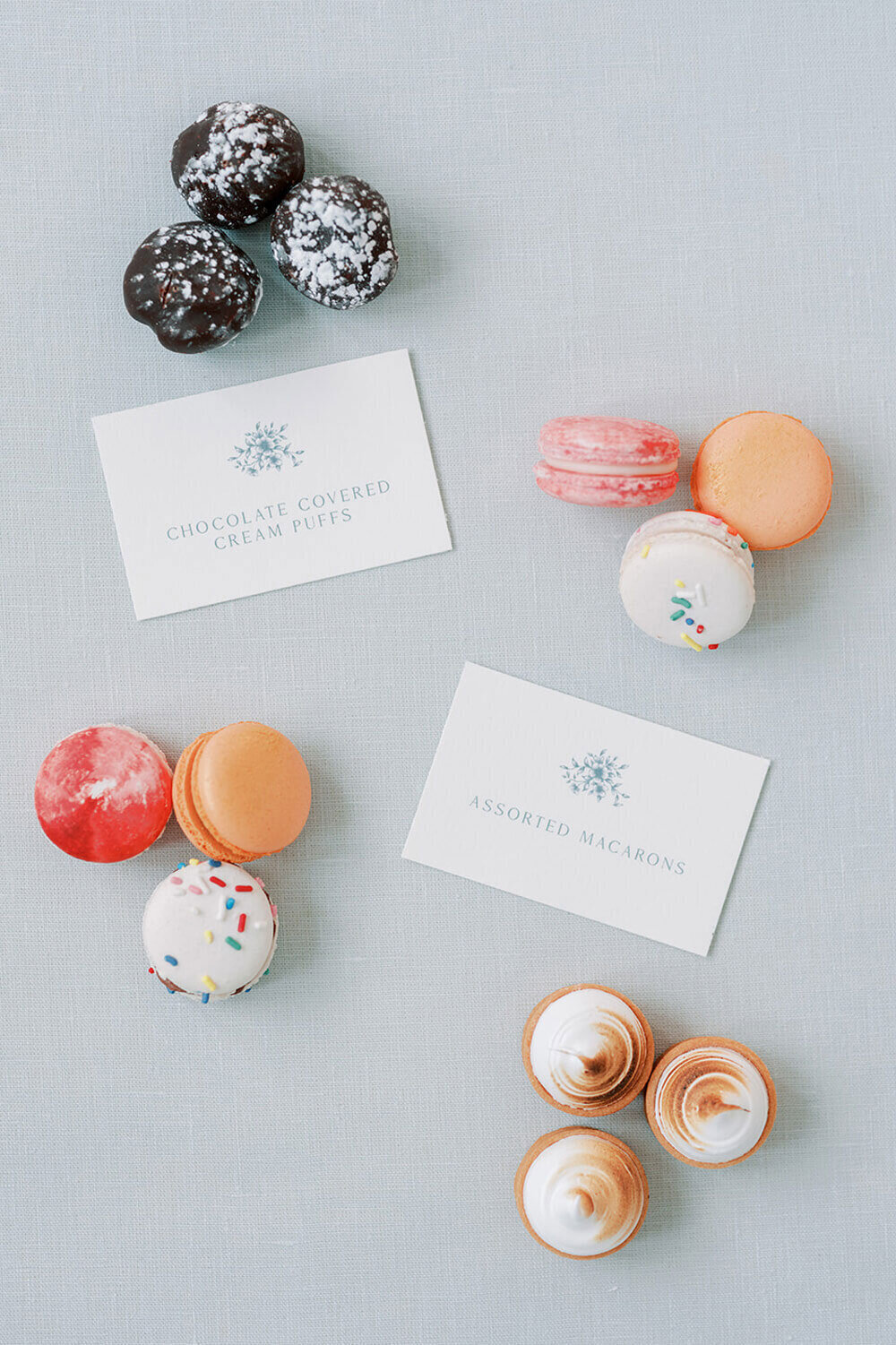 Assorted macarons, chocolate-covered cream puffs, and mini lemon tarts, paired with modern dessert signage on soft blue linen.