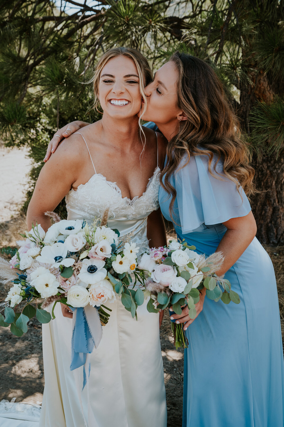 Bridesmaid kisses brides cheek while bride smiles holding bouquet.