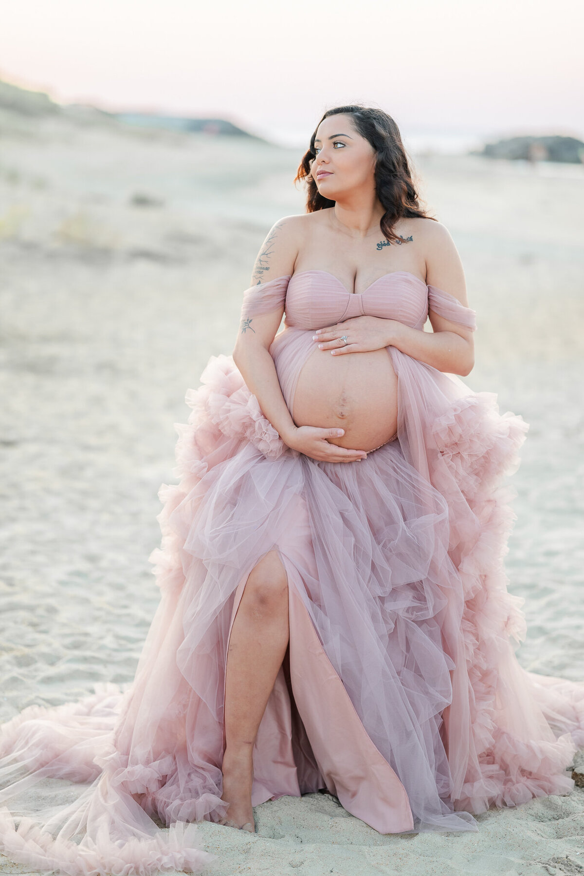 A pregnant woman holds her belly and looks off to her right. She wears a pink tulle gown during her beach maternity photos.