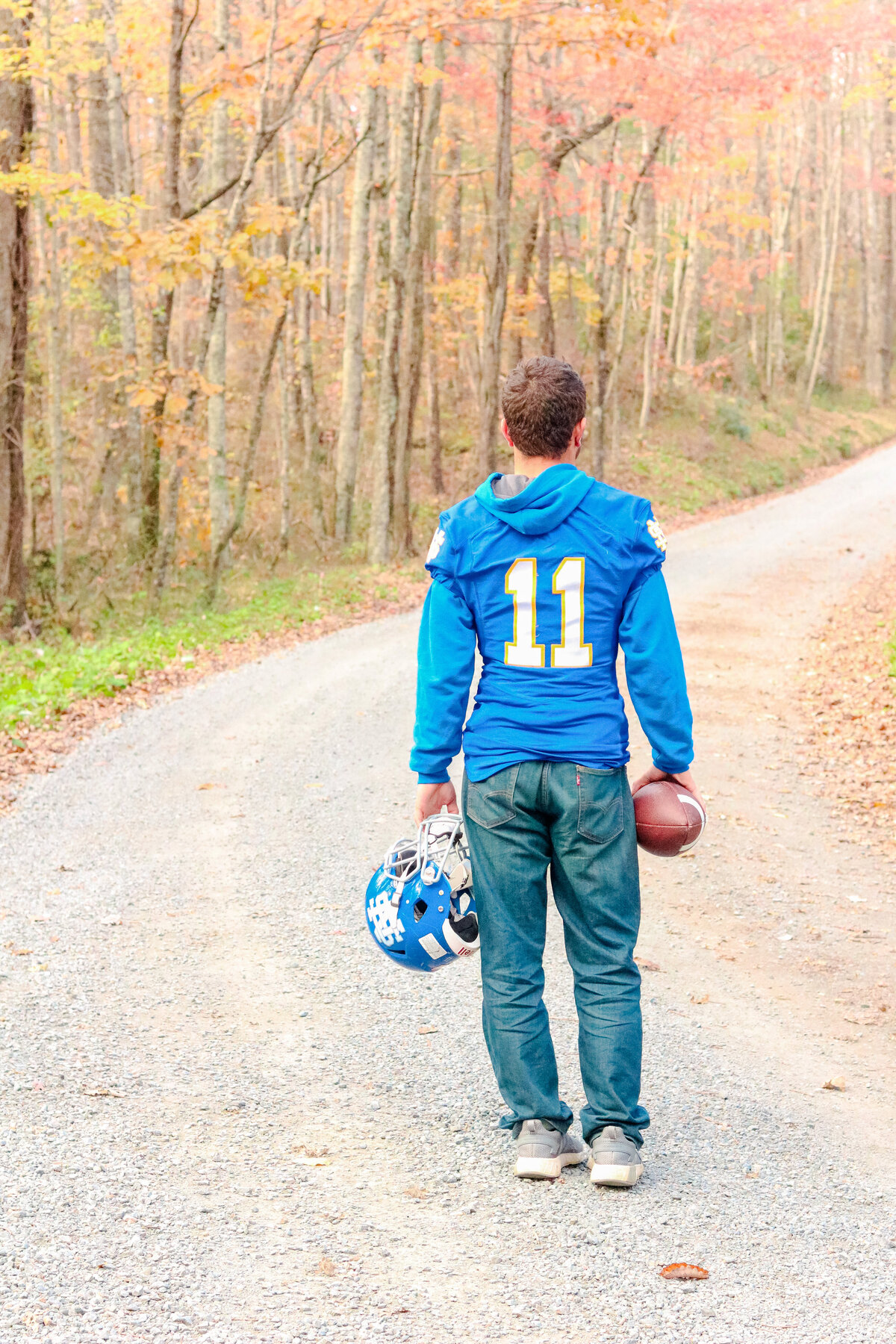 Senior guy standing on a dirt road in his football jersey with his ball and helmet