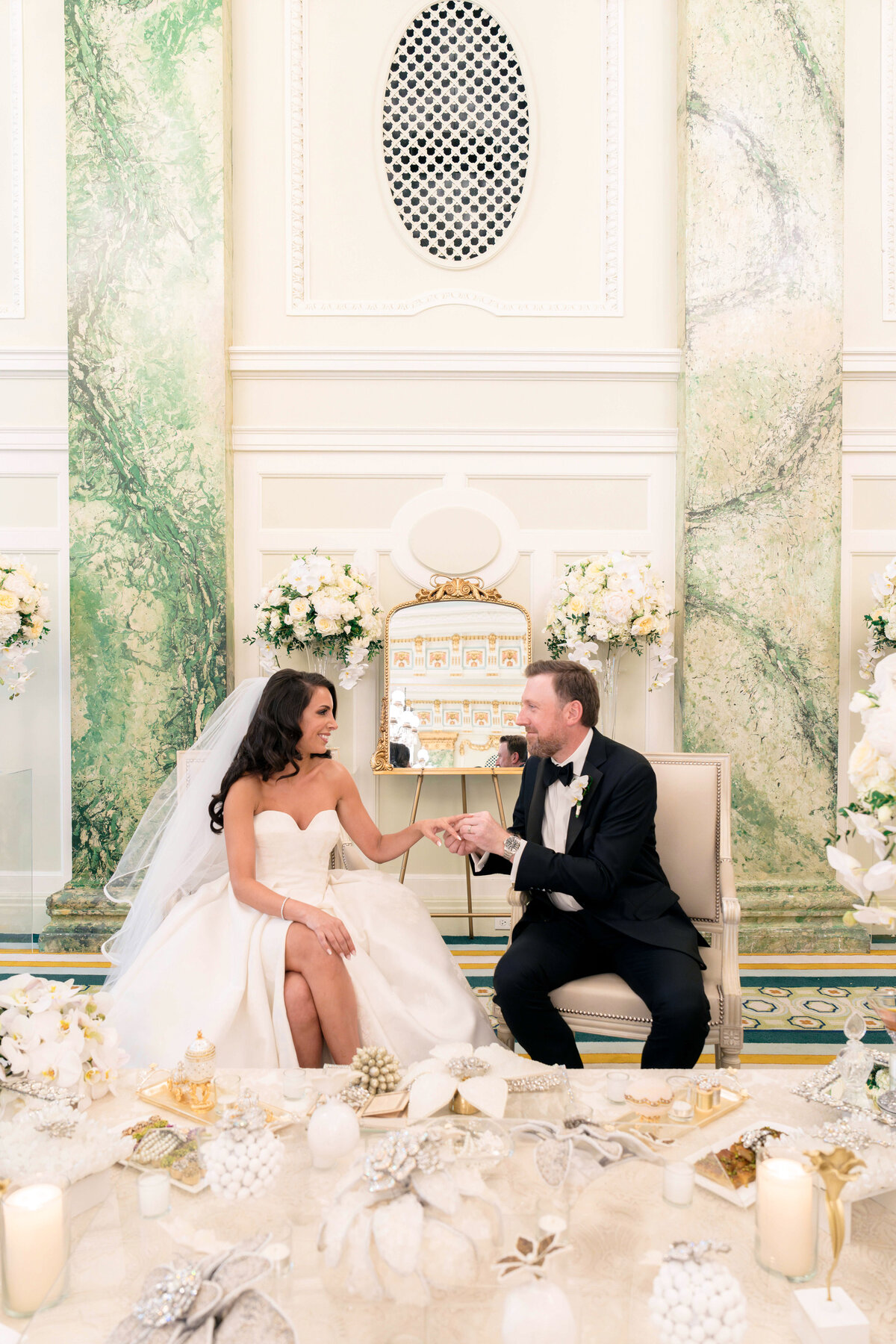 A bride and groom sit on elegant chairs, holding hands and smiling at each other. The bride wears a white gown and veil, while the groom is in a black suit. They are surrounded by white and gold decor with floral arrangements in a luxurious setting.