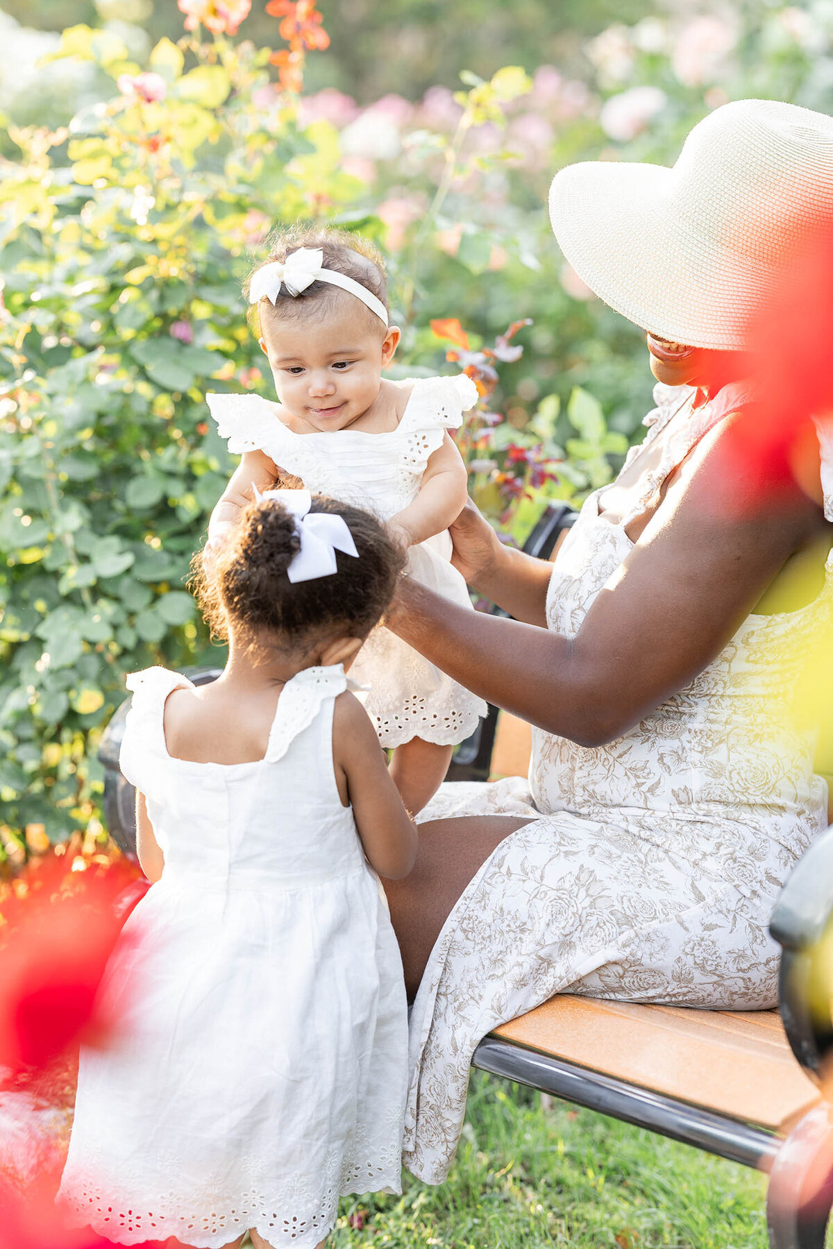 Baby girl in a while dress  plays with her toddler sister as mom holds baby girl up during a mommy and me session in a rose garden