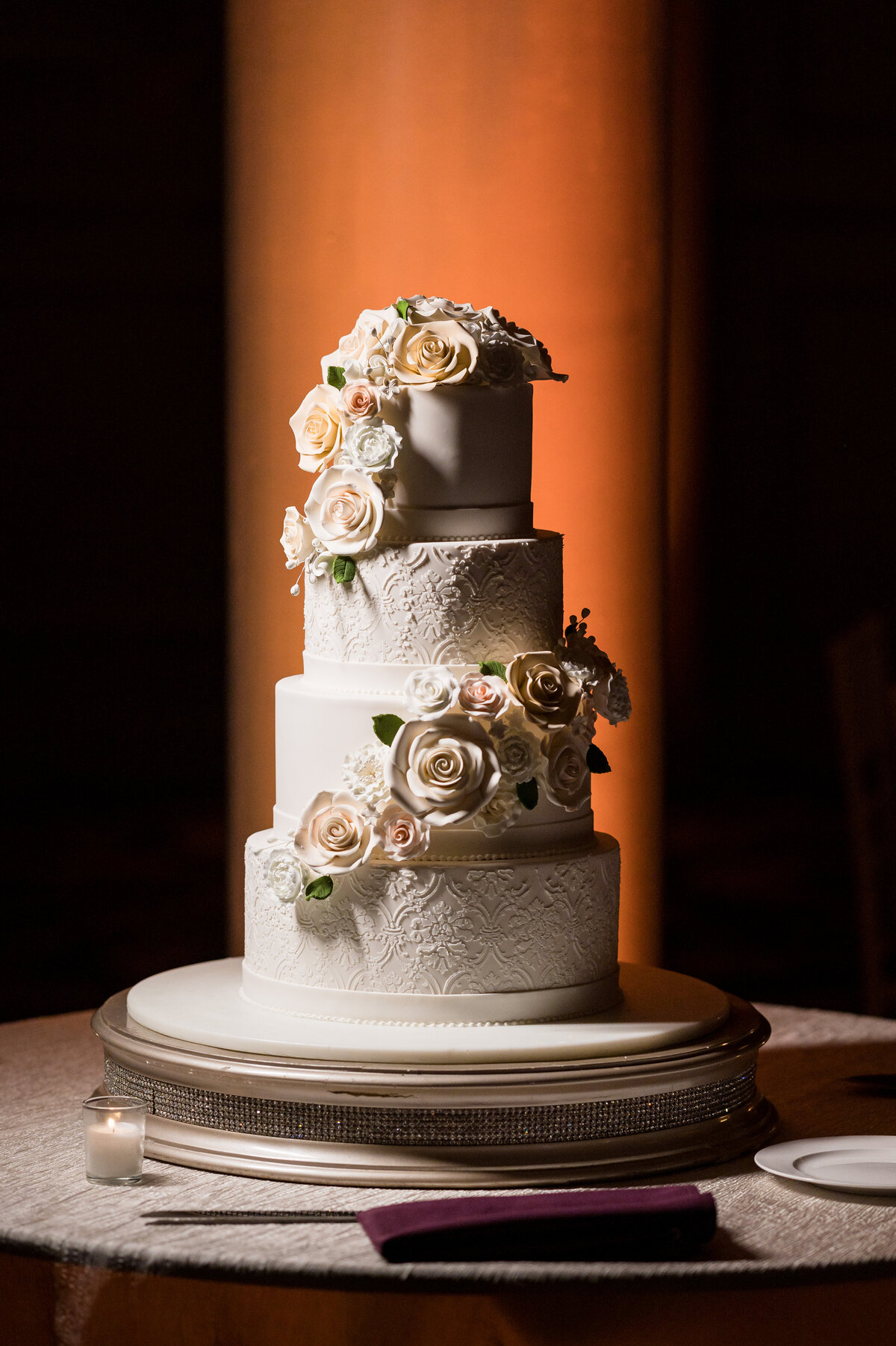 A three-tiered white wedding cake, adorned with intricate floral patterns and cascading cream and blush roses, graces a round table with a textured tablecloth. Perfect for capturing by a South Jersey wedding photographer.