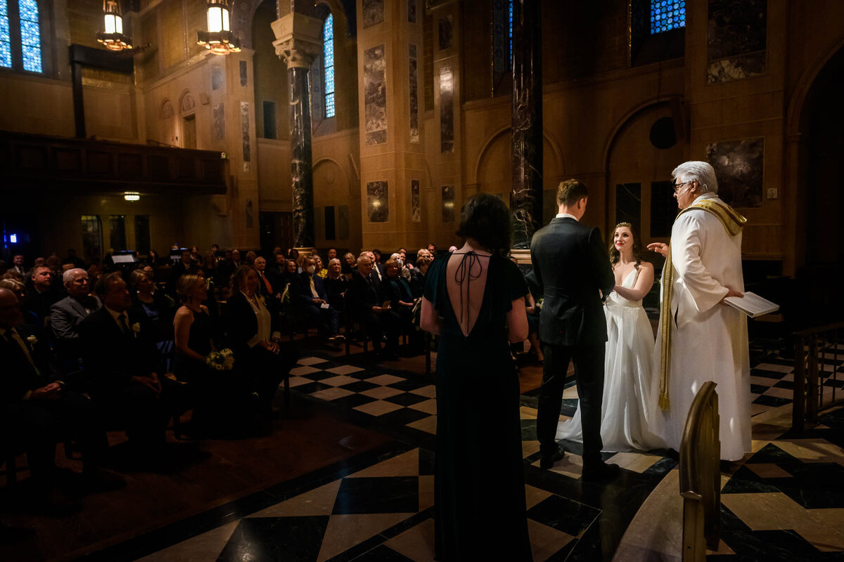 A couple stands together before their wedding guests at their ceremony in n a large, dramatically-lit chapel