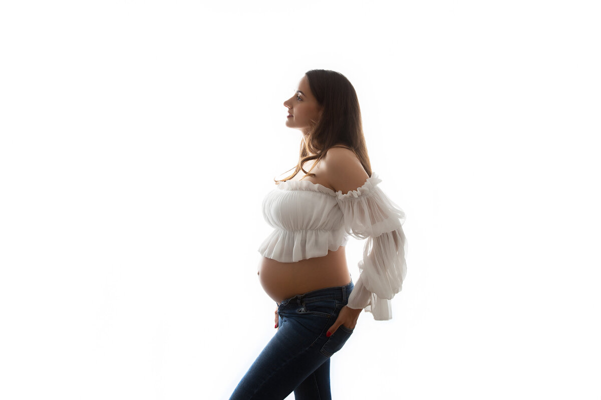 Pregnant woman posing  for a maternity portrait wearing blue jeans & a soft cropped top.