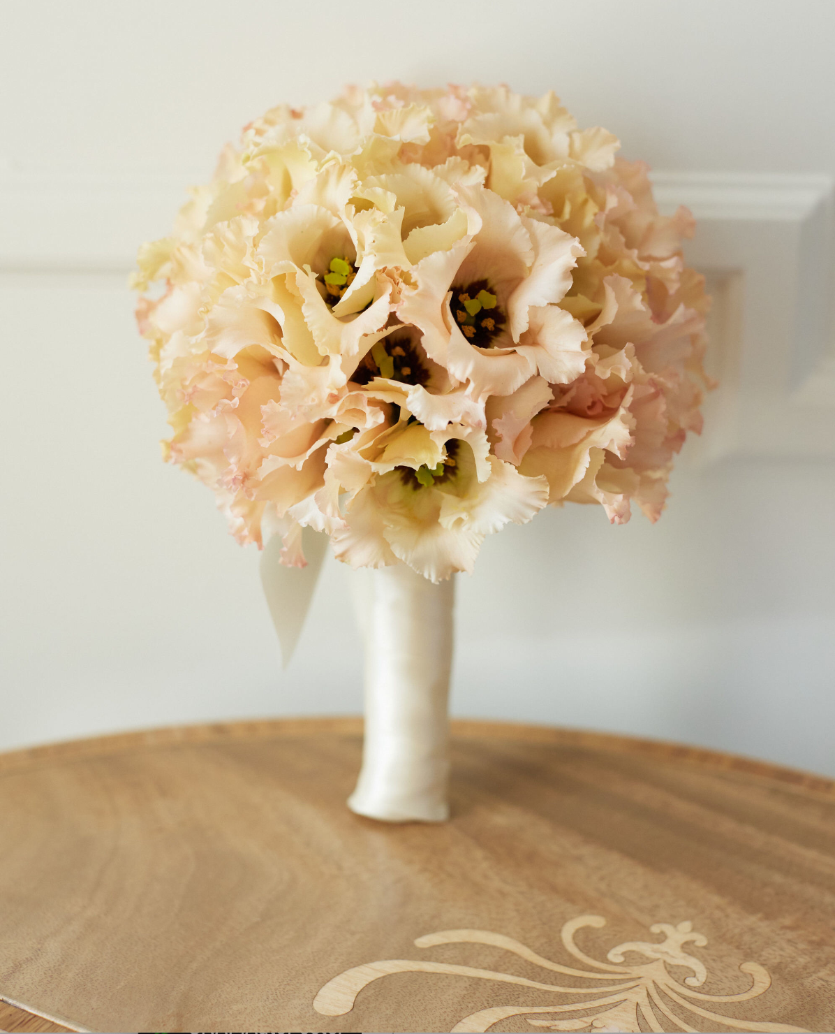 The photo was taken at The Willard Intercontinental in Washington D.C., known as one of the top wedding venues and hotels in DC, by photographer Shawn Connell. Videographers, Clayton Films, were also present to capture the special moments. In the image, a beautiful bouquet of pale peach and cream-colored flowers with ruffled petals is neatly arranged and wrapped in white satin ribbon. The bouquet is placed on a wooden table with a subtle floral design, adding a touch of elegance to the setting.