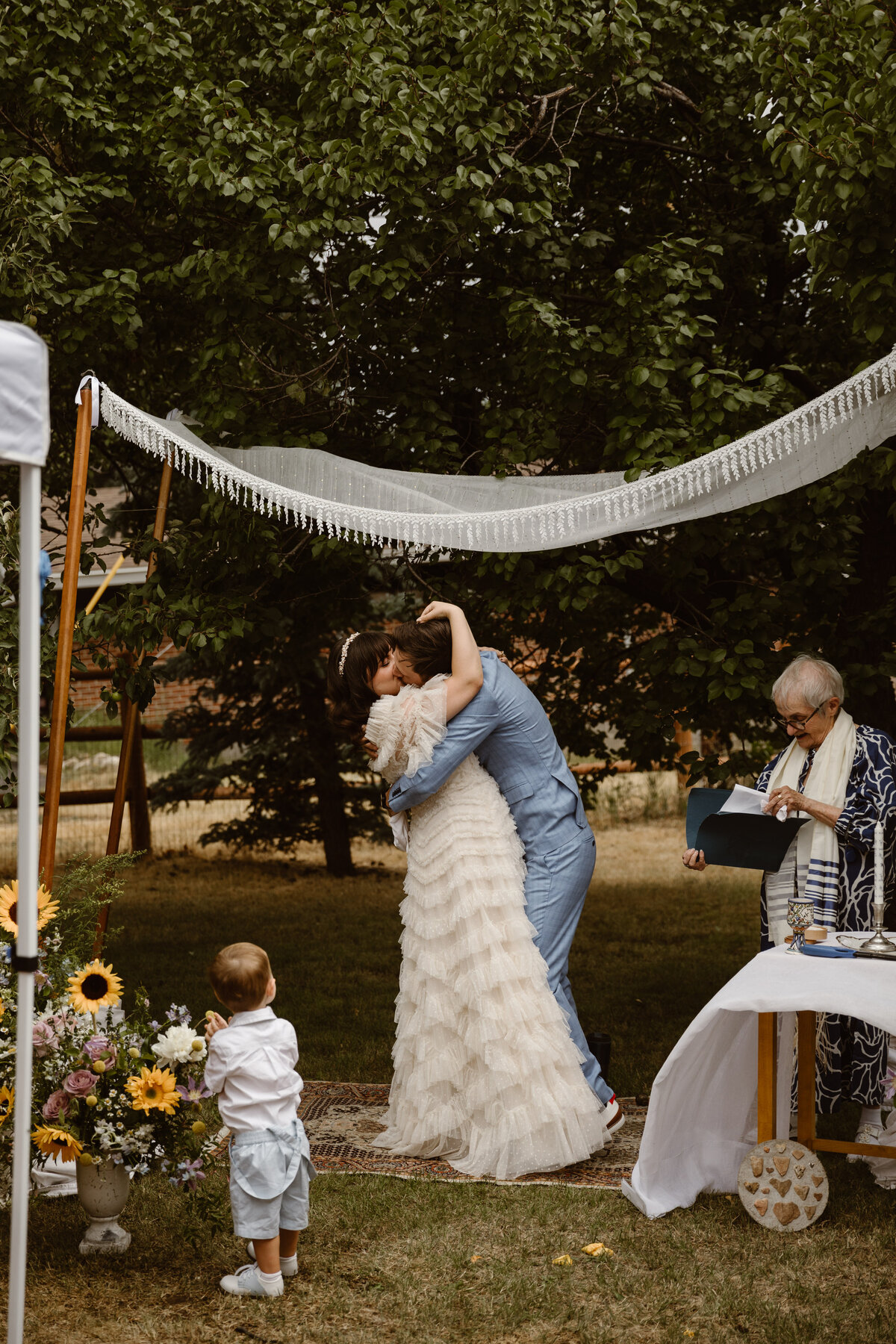 Bride and groom share their first kiss beneath a chuppah during an intimate backyard Jewish wedding ceremony, with their child looking on and the officiant reading beside them.