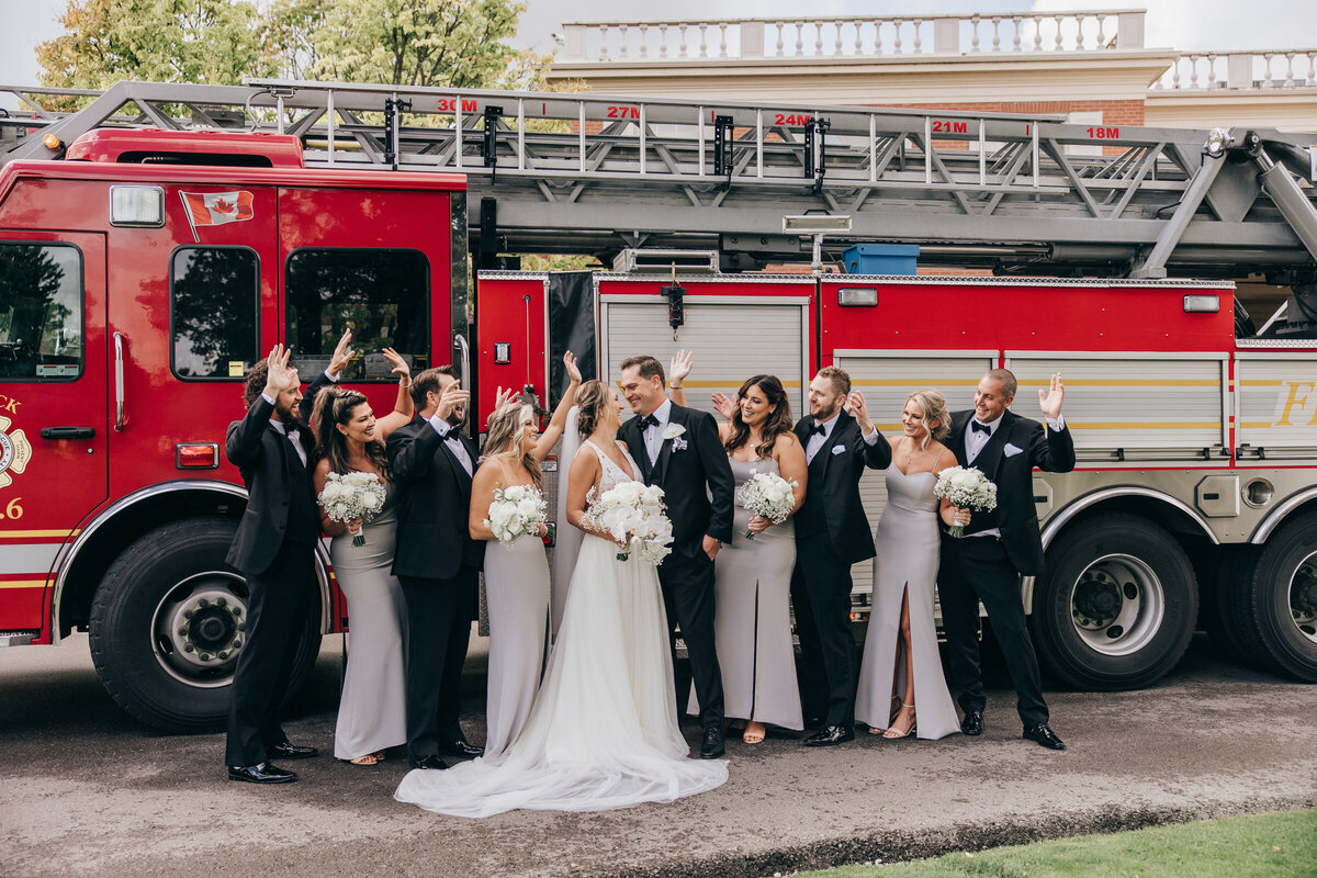 Large wedding party wearing black tie attire posing for photos in front of a red fire truck