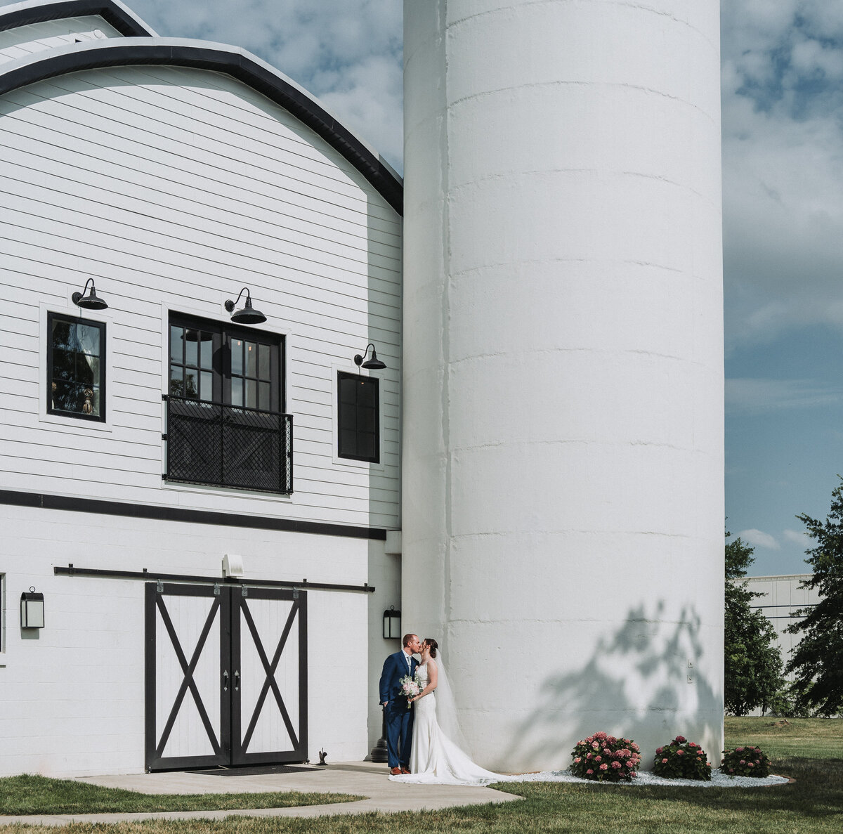 A newlywed couple shares a romantic kiss in front of the iconic white barn and silo at Sweeney Barn, a modern farm wedding venue in Manassas, Virginia, beautifully captured by Quinn Photography LLC.