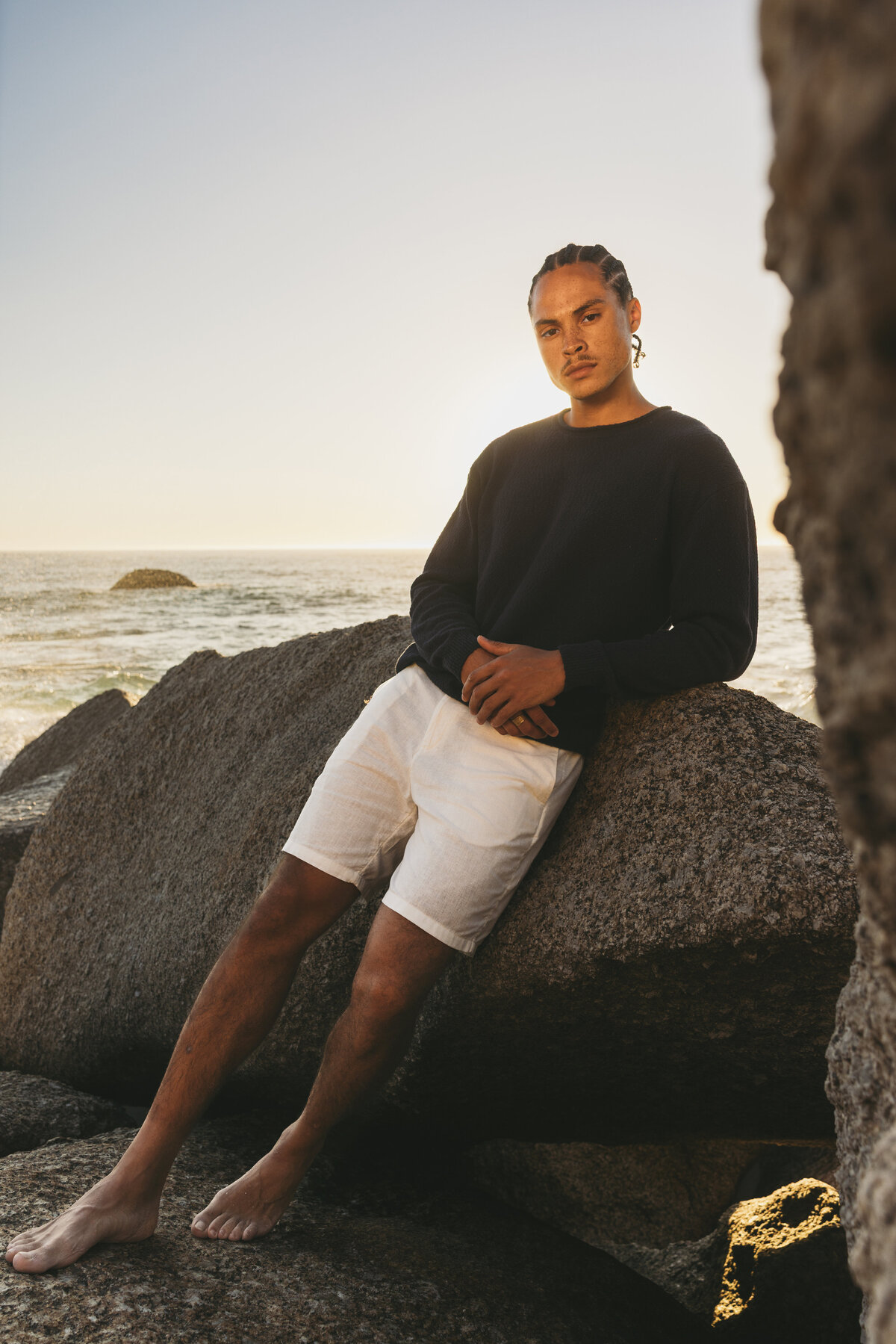 “Timothe leaning casually against coastal rocks during golden hour in Cape Town, wearing a black sweater and white shorts.