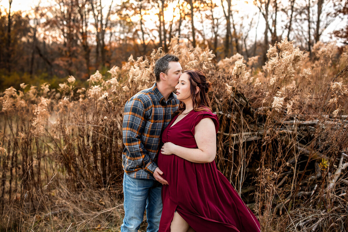 A man in a plaid shirt kisses a pregnant woman on the forehead. She wears a flowing red dress and rests her hand on her belly. They stand in a field with dried plants and trees in the background during sunset.