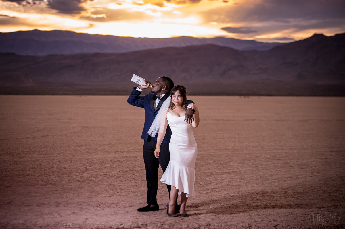 Groom tosses back bottle of champange while bride poses for camera with golden hour sun setting behind the mountains with purple skies las vegas elopement on the dry lake bed  at golden hour groom in blue suit jacket and black  pants  las vegas elopement eloping in vegas  las vegas wedding photographers las vegas wedding photography mk delacy photography