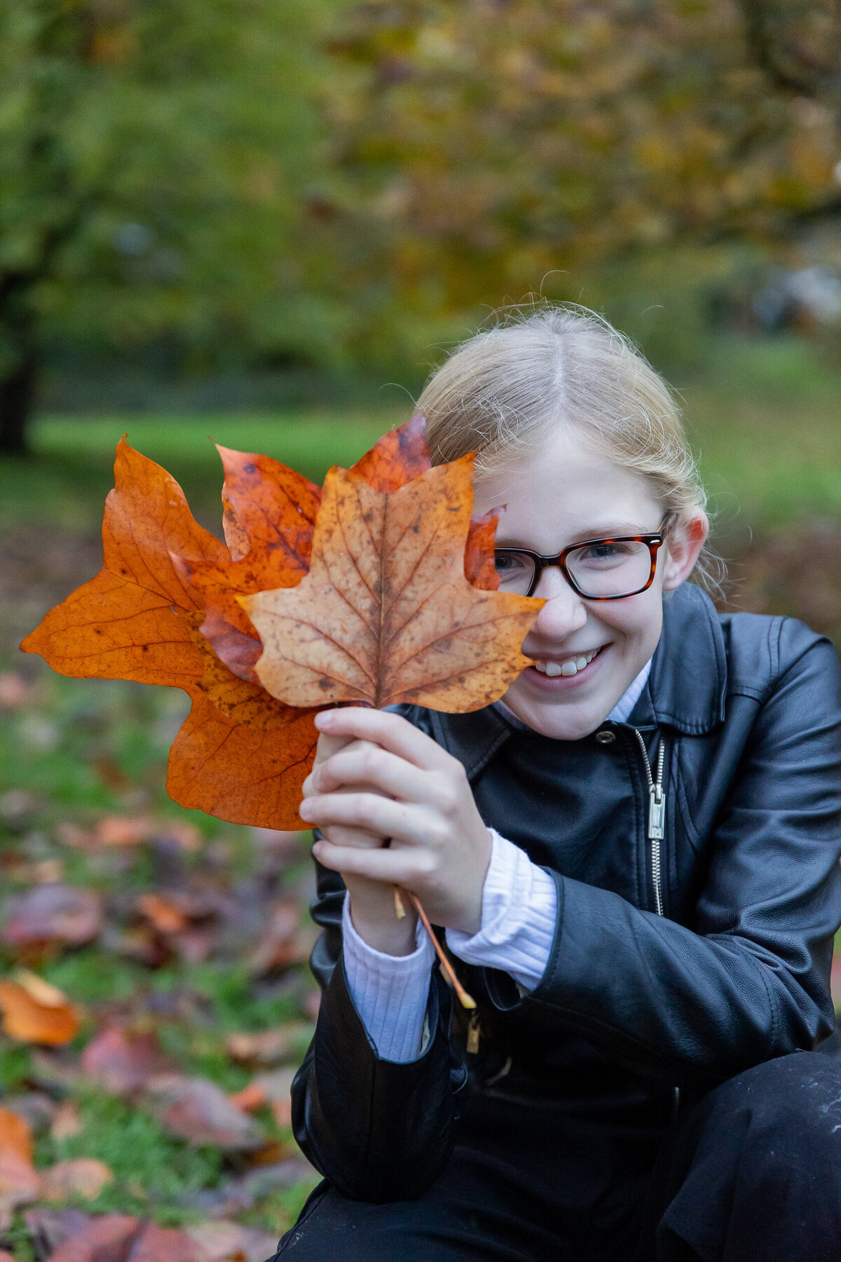 A young girl in a black jacket and glasses smiles while standing behind a younger boy in a blue sweater, both leaning on a stone wall in a grassy area.