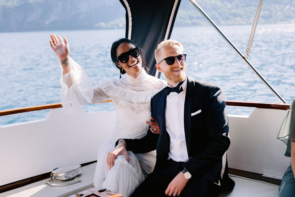 The bride is waving her hand while sitting with her groom inside a boat.
