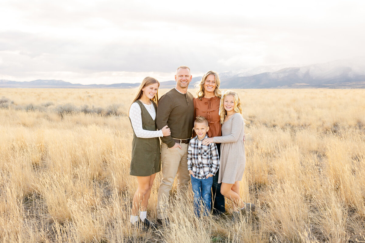 A family of five standing together in a golden field in Eagle Mountain, Utah, with mountains and soft clouds in the background. They are dressed in earthy tones, smiling warmly at the camera.