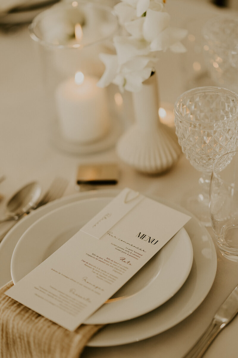 Assiettes blanches sur lesquelles sont déposées la carte du menu, entourées de couverts, verres et bougies. Ambiance blanche et beige douce et minimaliste. photo prise par Laura, photographe mariage en vendée.