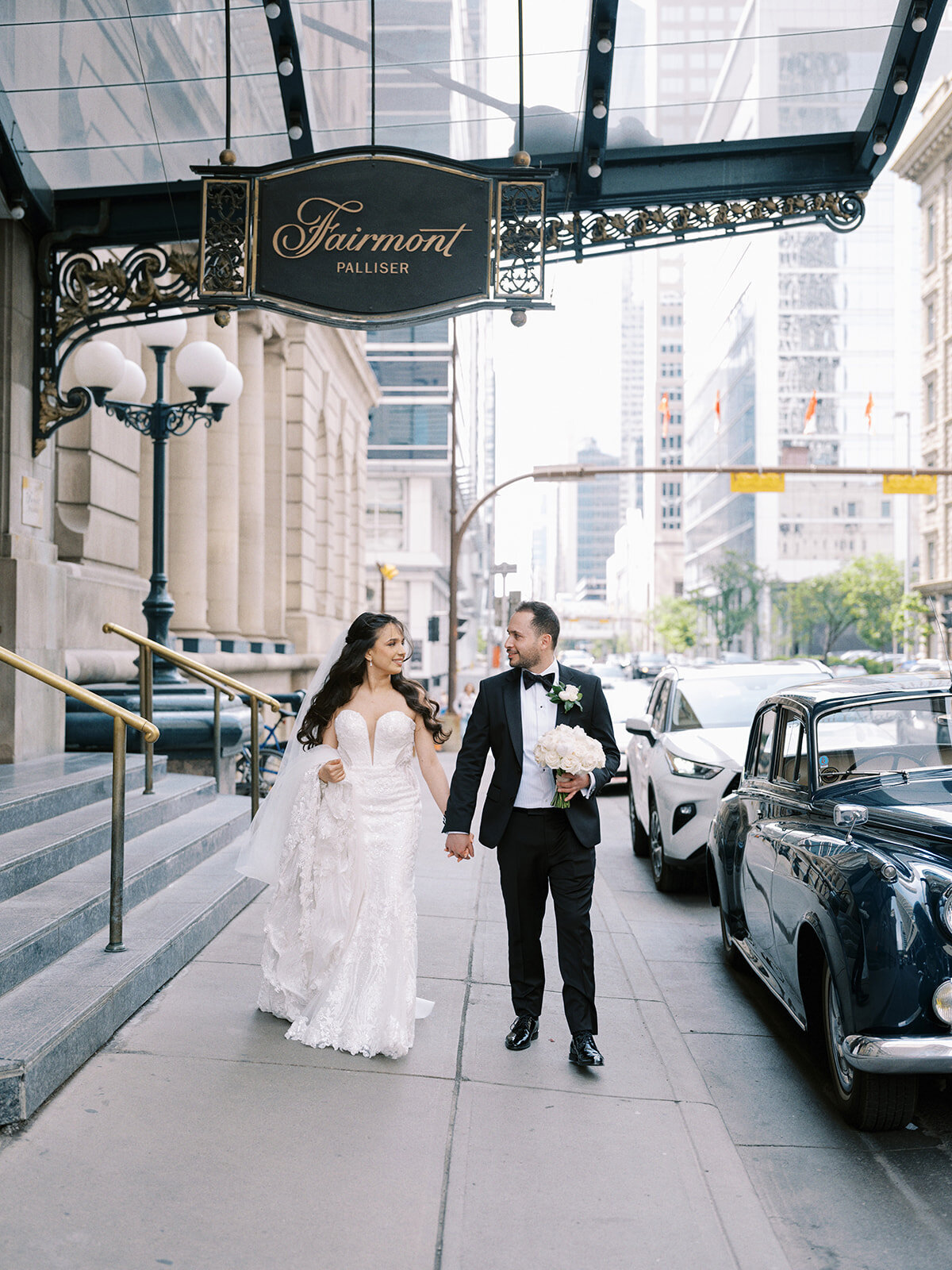 A bride and groom walk hand-in-hand outside the Fairmont Palliser hotel, epitomizing a classic Calgary wedding. They are dressed in wedding attire, with the groom holding a bouquet. Urban buildings and cars frame this beautiful moment at their Wedding at Fairmont Palliser Calgary.