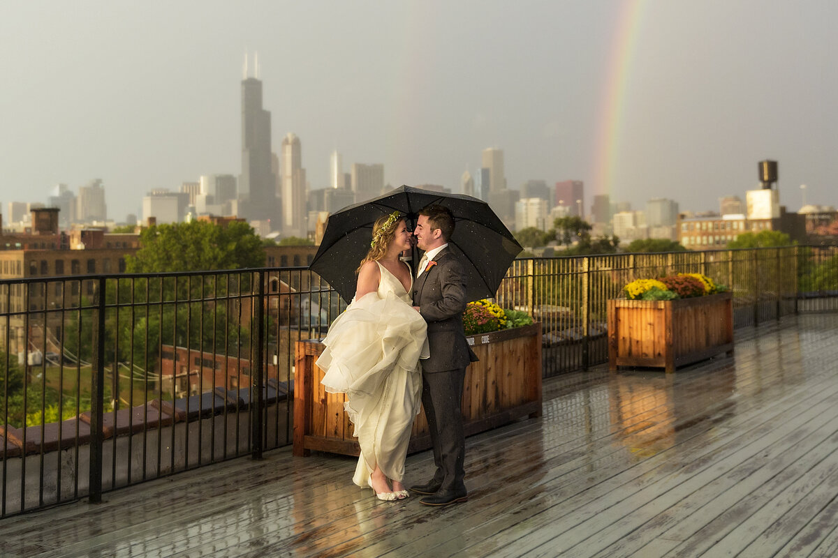 A bride and groom under an umbrella with a rainbow on the lacuna lofts rooftop.
