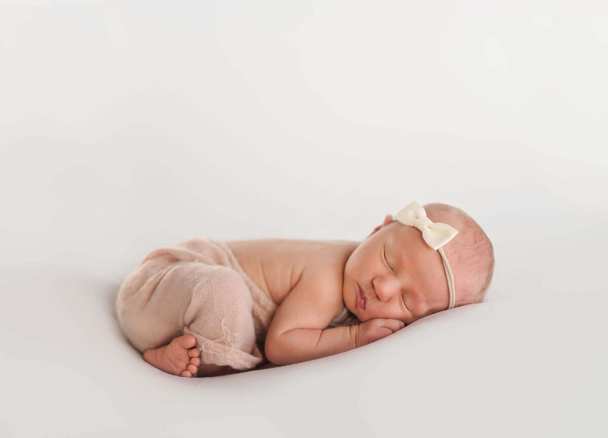 A newborn baby girl  with a pink headband on sleeps peacefully on a white blanket during her session with an Asheville Newborn Photographer