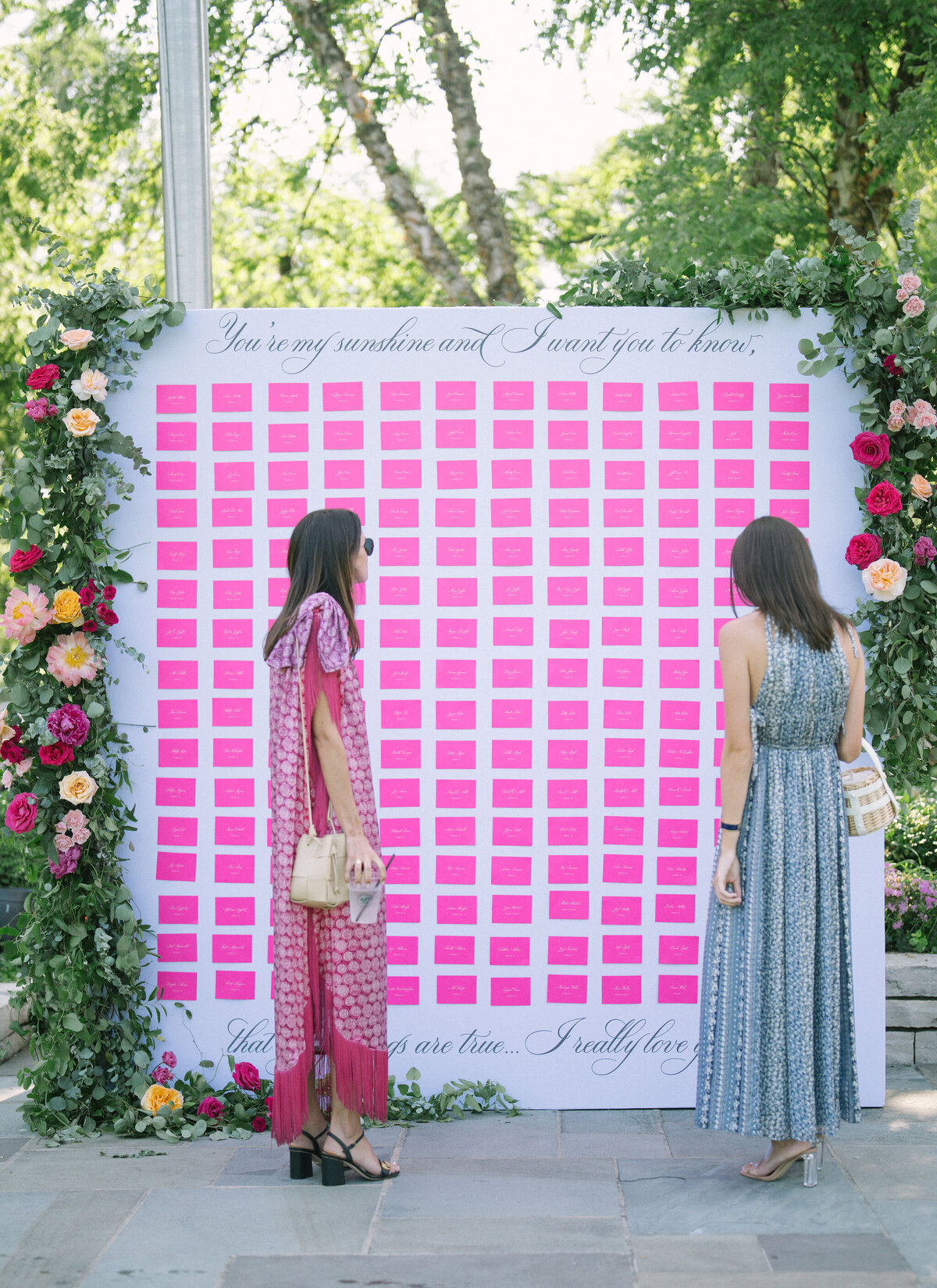 Two guests admiring a pink seating chart at a floral photo wall at Saddle Country Club.