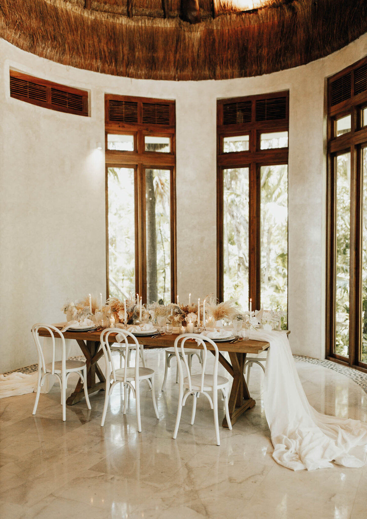 A rustic indoor setting with a wooden dining table decorated with beige and white candles, dried floral arrangements, and a white table runner. The room has large windows and white chairs, creating a warm, elegant atmosphere.