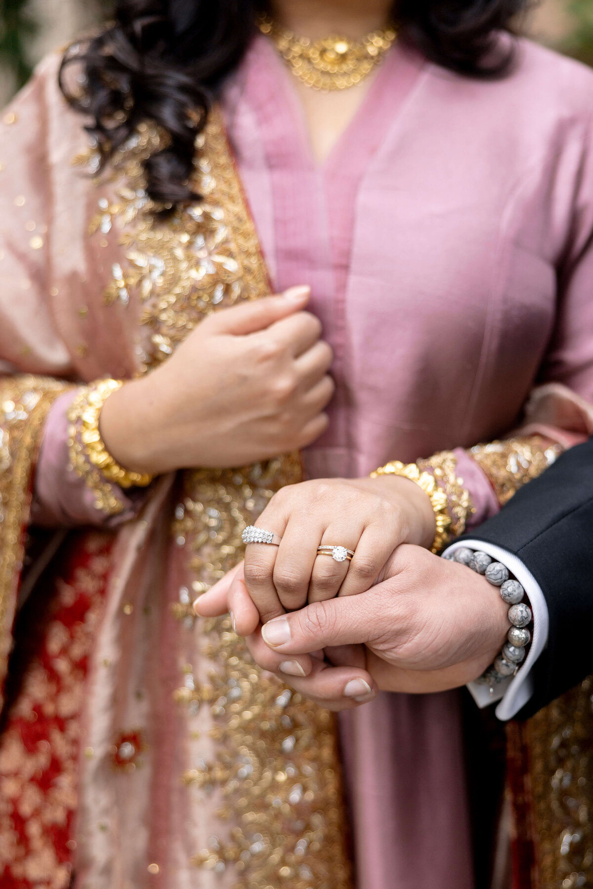 A couple holds hands, showcasing rings. The woman wears a pink and gold embellished traditional outfit with gold jewelry, while the man wears a black suit with a beaded bracelet. The image focuses on their intertwined hands.