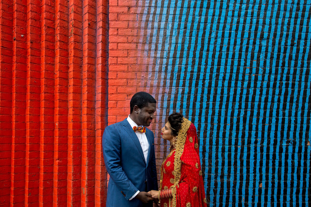 A couple holding hands and looking at each other in front of a colorful wall.