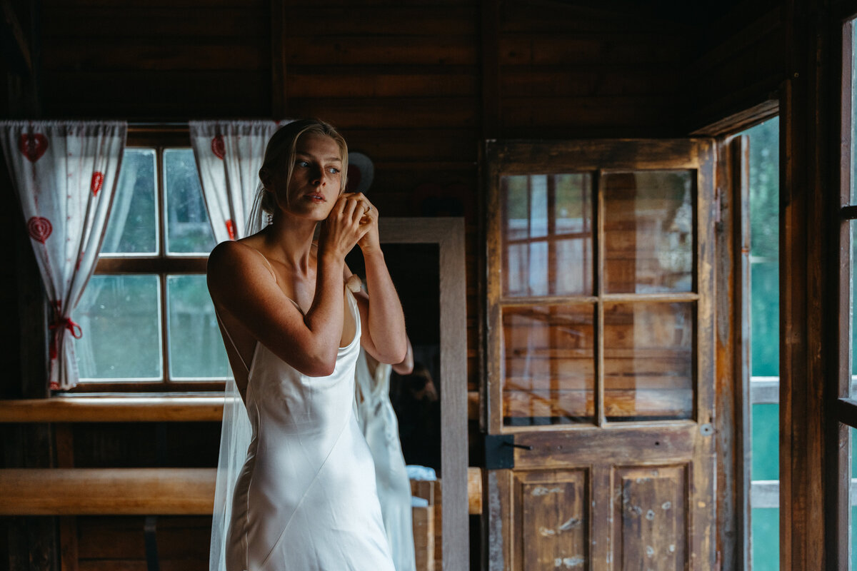 Bride in satin dress putting on earrings by wooden door