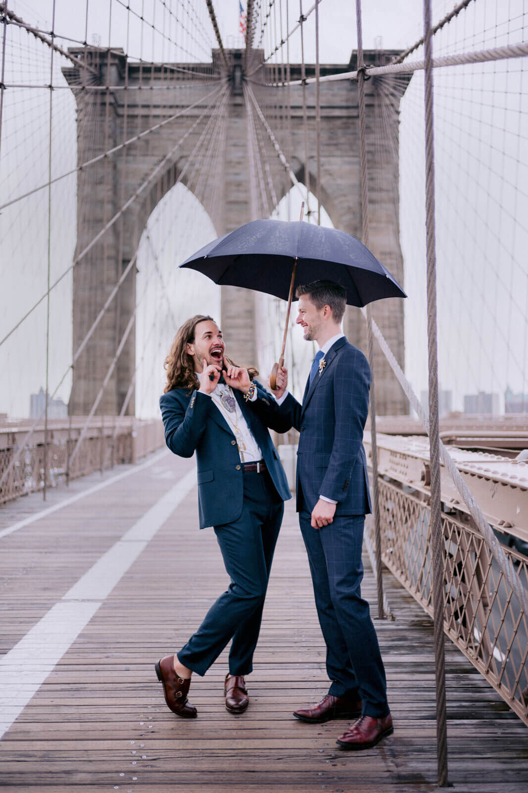 The two grooms are happily chatting under an umbrella on the Brooklyn Bridge. NYC City Hall Elopement Image by Jenny Fu Studio