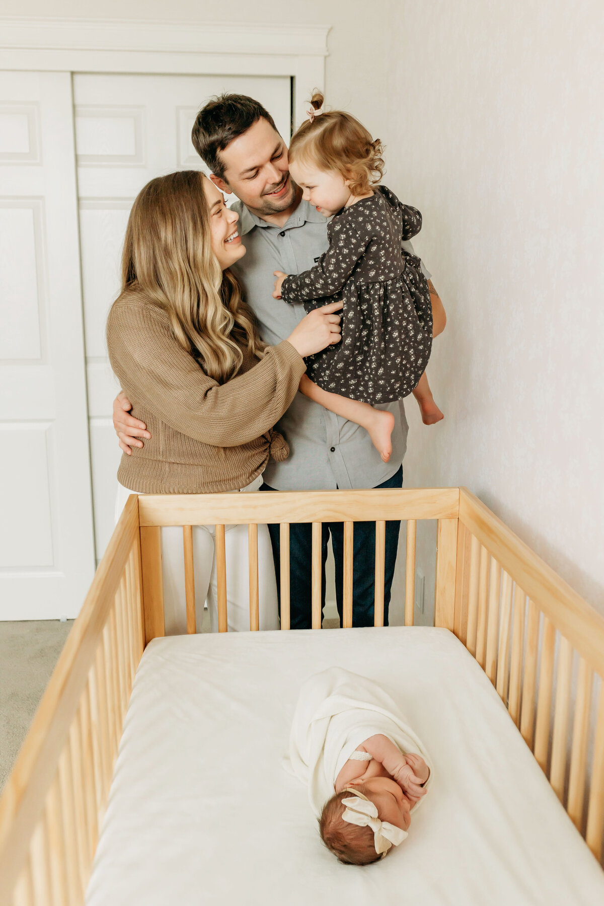 A mom, dad, and toddler stand at the foot of a crib within a nursery and admire the newborn baby inside the crib