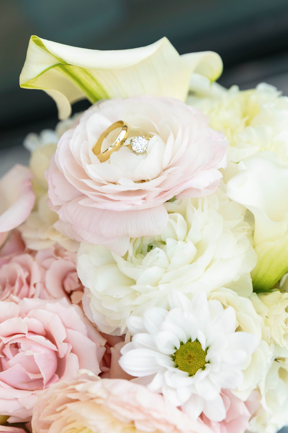 Close-up shot of wedding rings displayed on soft pastel flowers, including roses and ranunculus, showcasing gold bands and diamond engagement ring in romantic wedding detail photography.