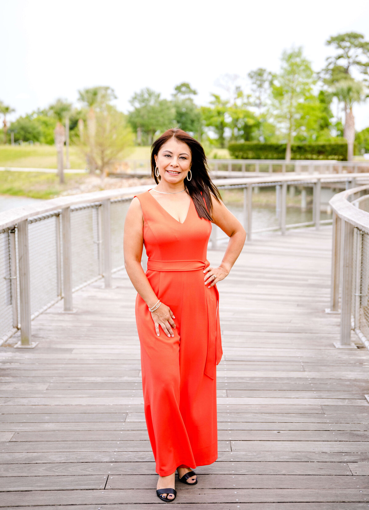 Brand photographer near me photographs woman in a bright red jumpsuit standing on a dock that goes over a small lake with lush palm trees and tropical plants behind her