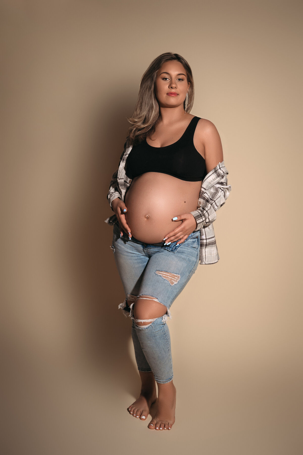 blonde pregnant woman wearing ripped jeans and an open button down plaid shirt holding her baby bump while looking at the camera