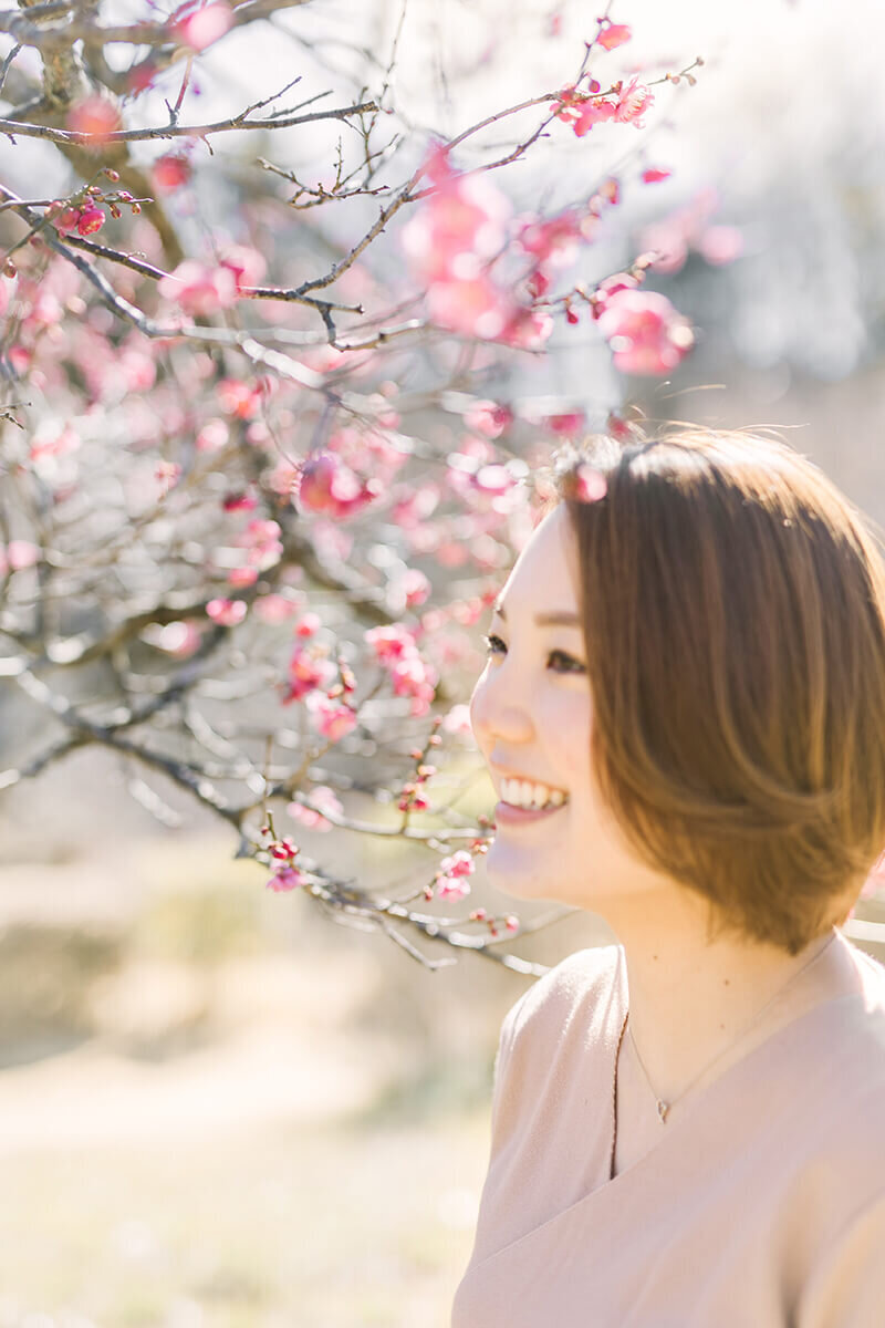 光輝いているピンクの梅と、その前でニッコリ笑っている女性の美しい写真