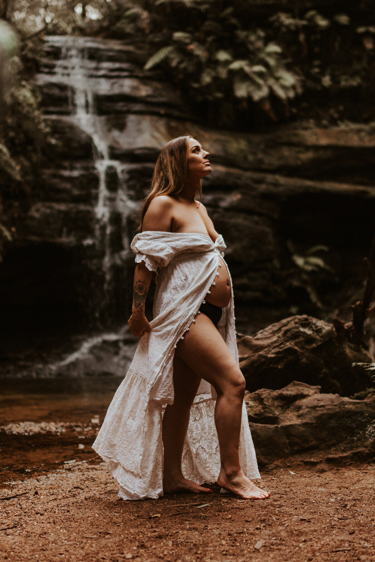 Pregnant woman in a white boho dress standing near a Sydney waterfall during an outdoor maternity photography session, embracing nature and motherhood