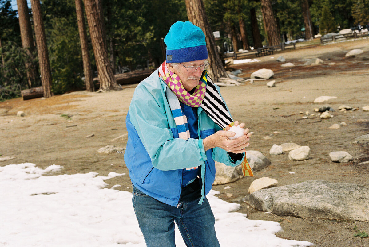 Alt text:
Old man playing with a snowball captured on film by Kate Winters Photography, showcasing a joyful and candid moment in an authentic, documentary style.